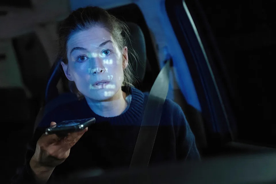 Rosamund Pike in a scene from "Hallow Road." A blond woman sits in a car at night and holds her smartphone up.