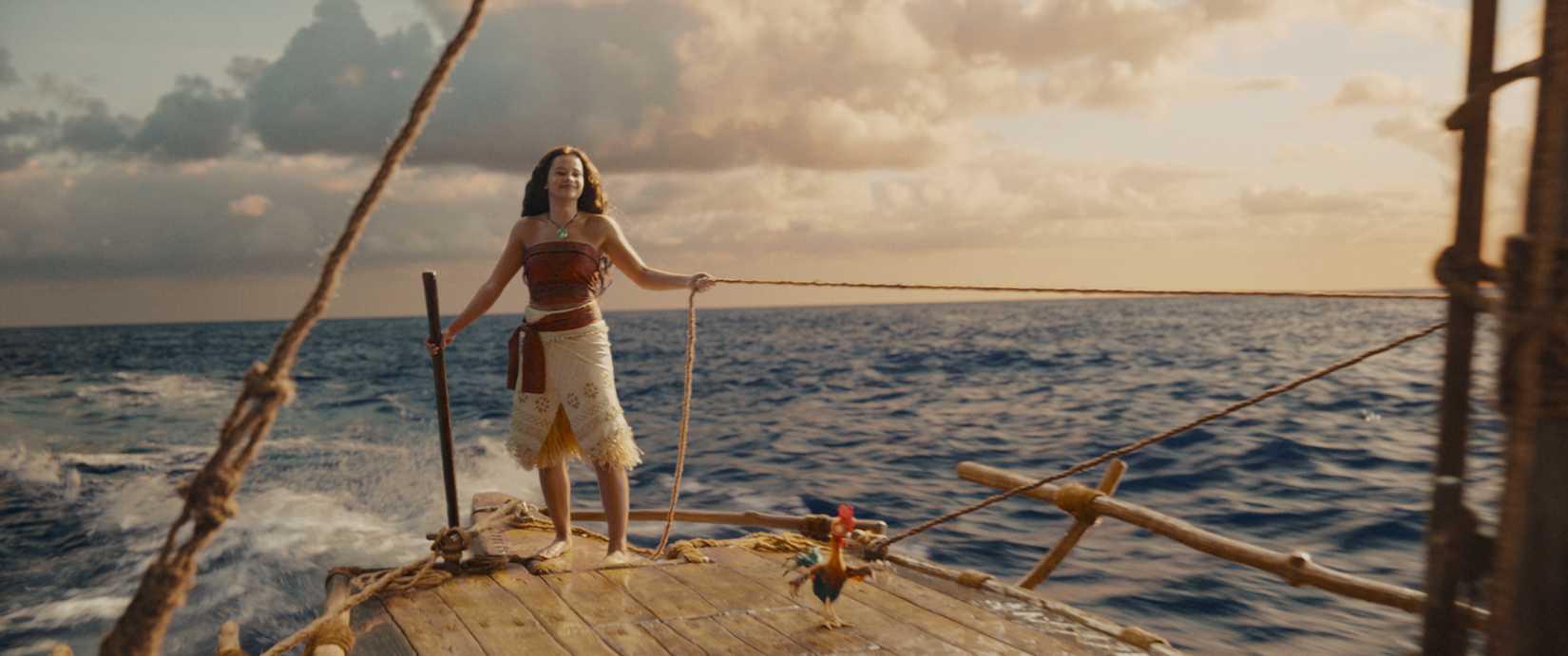Moana (Catherine Laga'aia), a Pacific Islander teenager, stands on the deck of a ship at sea with her (animated) rooster Hei-hei in the live-action Moana