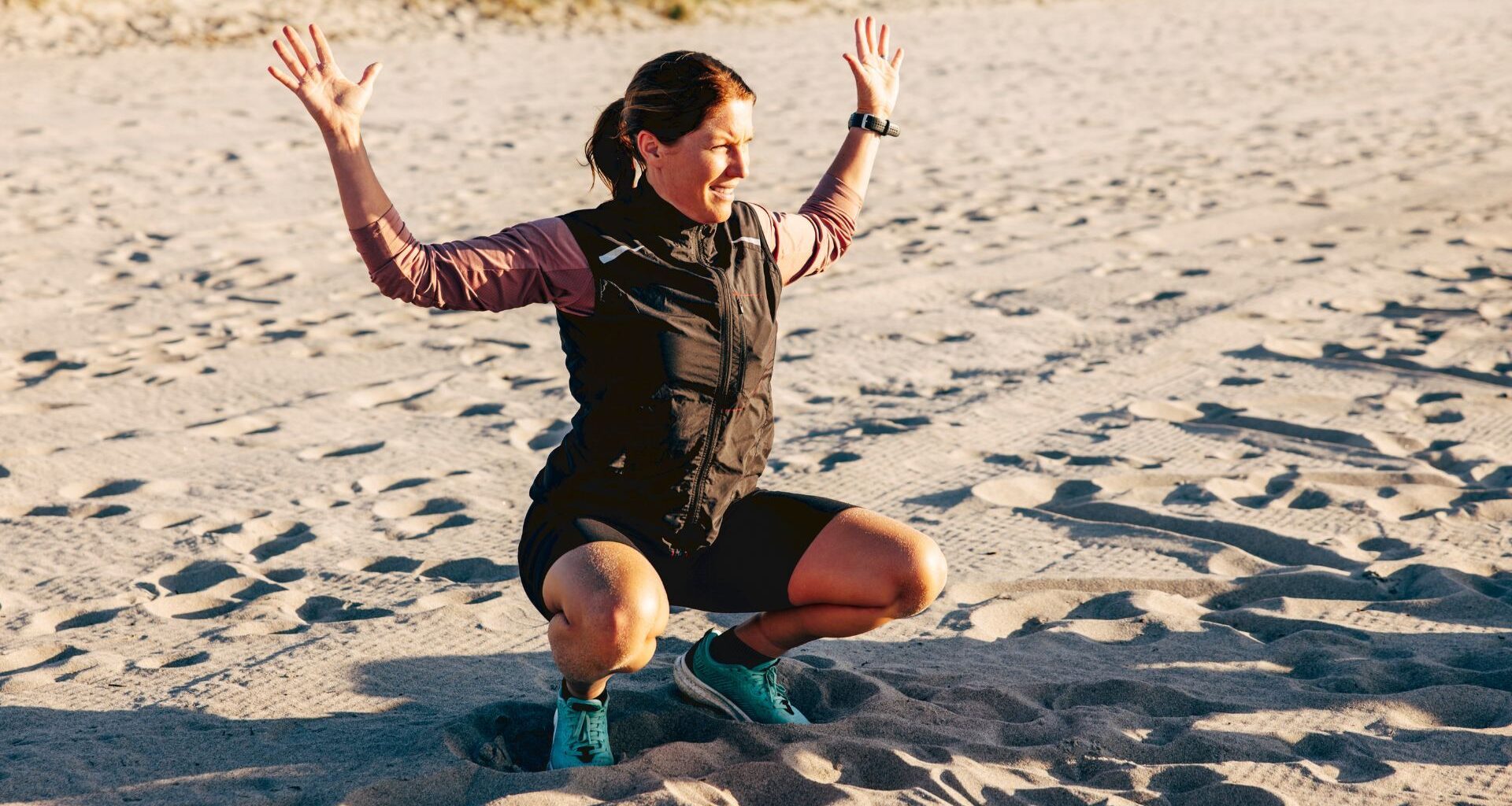 Woman at the bottom of a squat with hands in the air on the beach