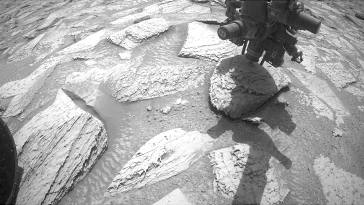A black and white still of NASA's Curiosity Mars rover just lifting its drill arm with a rock stuck to it
