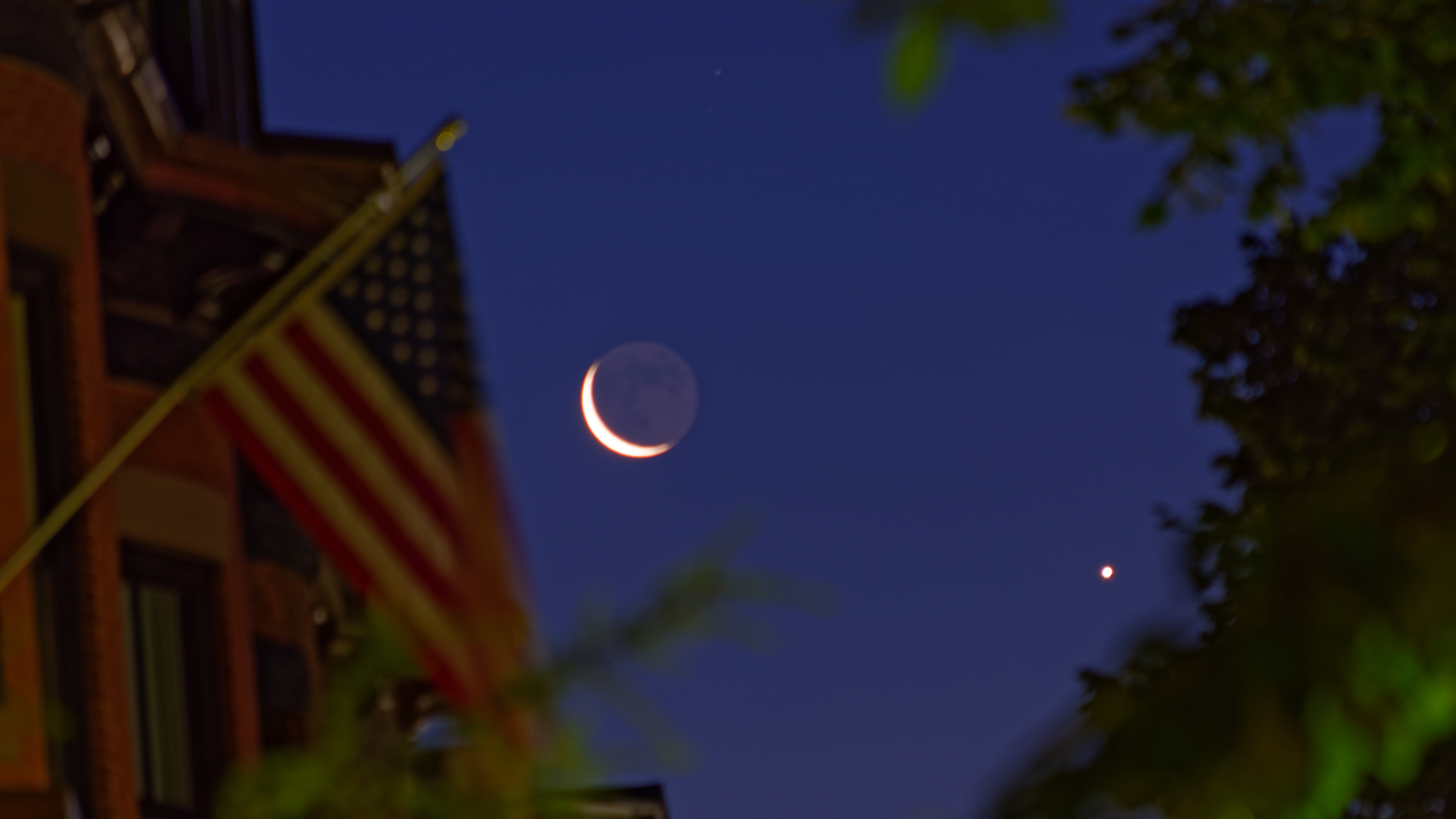 Rising moon in conjunction with Venus is framed on the left by brick townhouses with wooden dormers and an unfurled American flag in Boston's historic South End. Leafy treetops complete the framing on the right. The photo was taken at the start of nautical twilight.