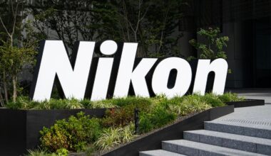 Large, white "Nikon" sign stands amid greenery outside a modern building, near concrete steps and surrounded by trees and bushes.