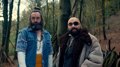 Two men, one in a blue denim jacket and the other with a big grey coat and an eye patch. They are looking into the camera stood in a wood