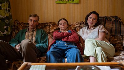 A man and a woman with a younger girl sat on a brown 1970s style sofa
