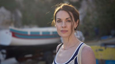 Jessica Raine in character as Zoe. She looks directly to the camera. Warm sunlight makes her hair glow. The background is blurred but a boat on a trailer is visible. 