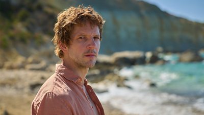 Damien Molony in character as Dan. Stood on a sandy beach in the sunshine, he wears an open shirt and watches something offscreen. 