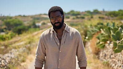 Nicholas Pinnock in character as Solomon. Wearing sunglasses and a shirt, he walks along a road in the sunshine. A row of cacti line the roadside. 