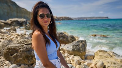 Leila Farzad in character as Nat. Wearing sunglasses, she smiles and looks directly to the camera. She’s sat on rocks beside the sea. 