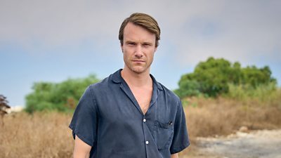 Hugh Skinner in character as Jacob. Wearing a short sleeved, blue shirt, he stands in the countryside and looks directly to the camera. 