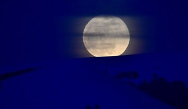 A hazy full moon wises over a snow covered hill at night.
