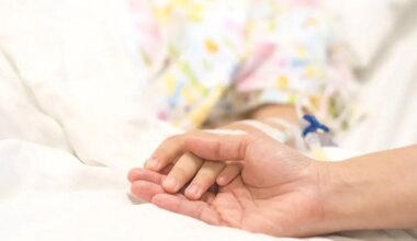 A close-up of an adult gently holding a child’s hand in a hospital bed. The child is wearing a colorful hospital gown and has a medical IV line attached, conveying comfort, care, and support during treatment.