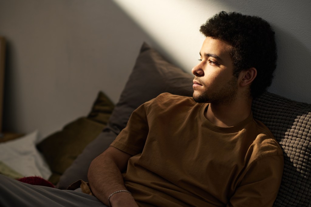 A sad teenage boy sitting alone on a bed in his room, looking lost in thought.