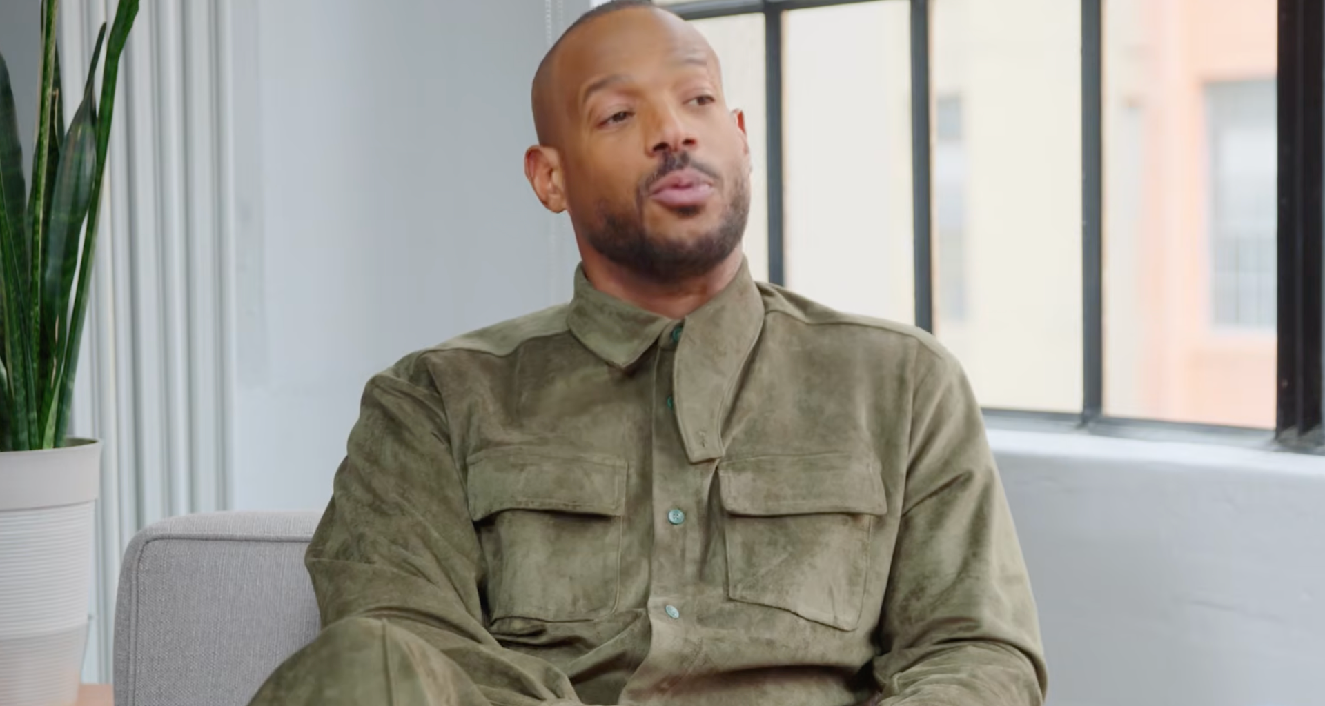 Marlon Wayans in a green outfit sits on a couch, speaking.