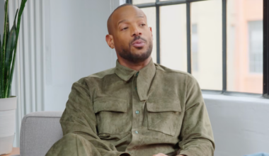 Marlon Wayans in a green outfit sits on a couch, speaking.