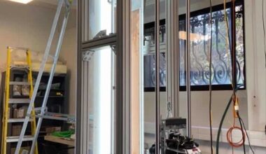 Photo of the experimental apparatus in the laboratory, showing a tall, well-lit transparent box supported by a metal frame. A camera is mounted on a vertical track next to the box and there are ladders in the background.