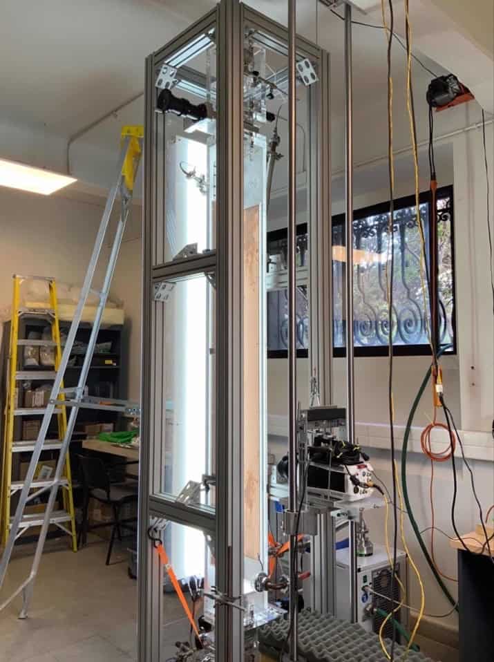 Photo of the experimental apparatus in the laboratory, showing a tall, well-lit transparent box supported by a metal frame. A camera is mounted on a vertical track next to the box and there are ladders in the background.