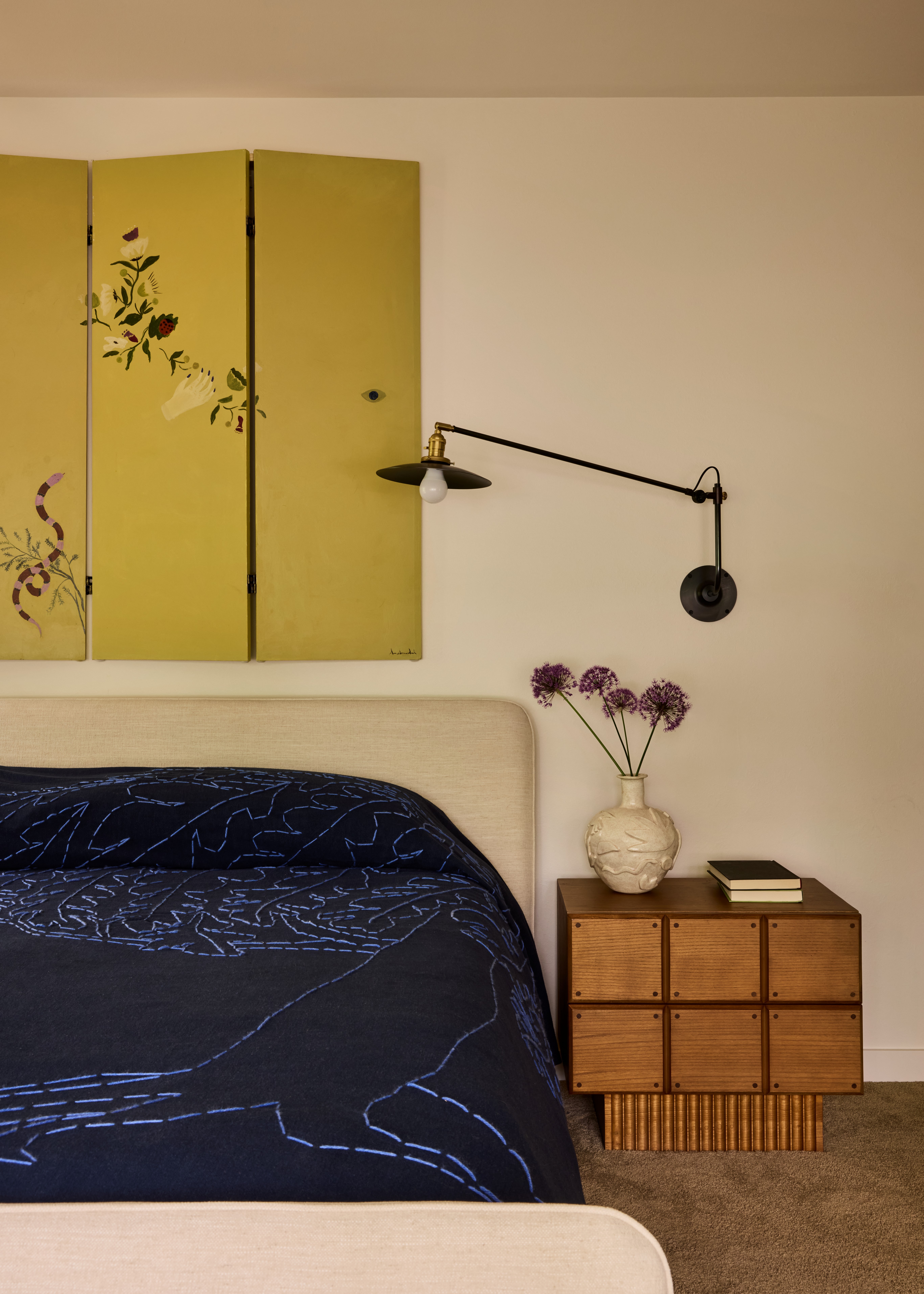 A bedroom with a separee hung as a tapestry above the headboard in yellow-tinted wood with florals, a cream headboard and blue bedsheets with handwoven motifs, a brown carpet and wooden furniture.