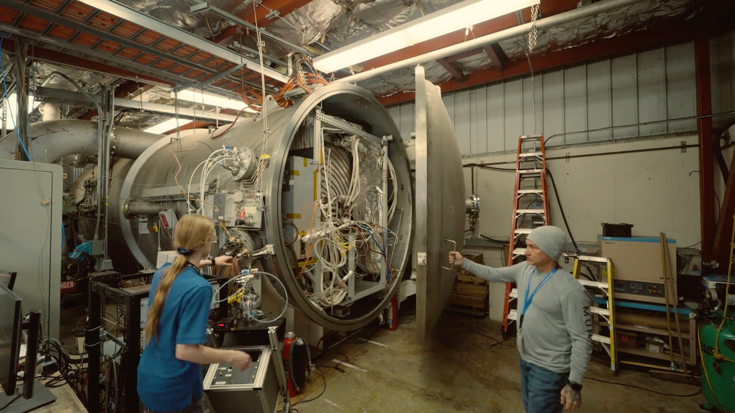 Two people stand in front of a giant silvery cylinder that's horizontally placed. One person is opening the dome of the cylinder and there are tons of wires within.