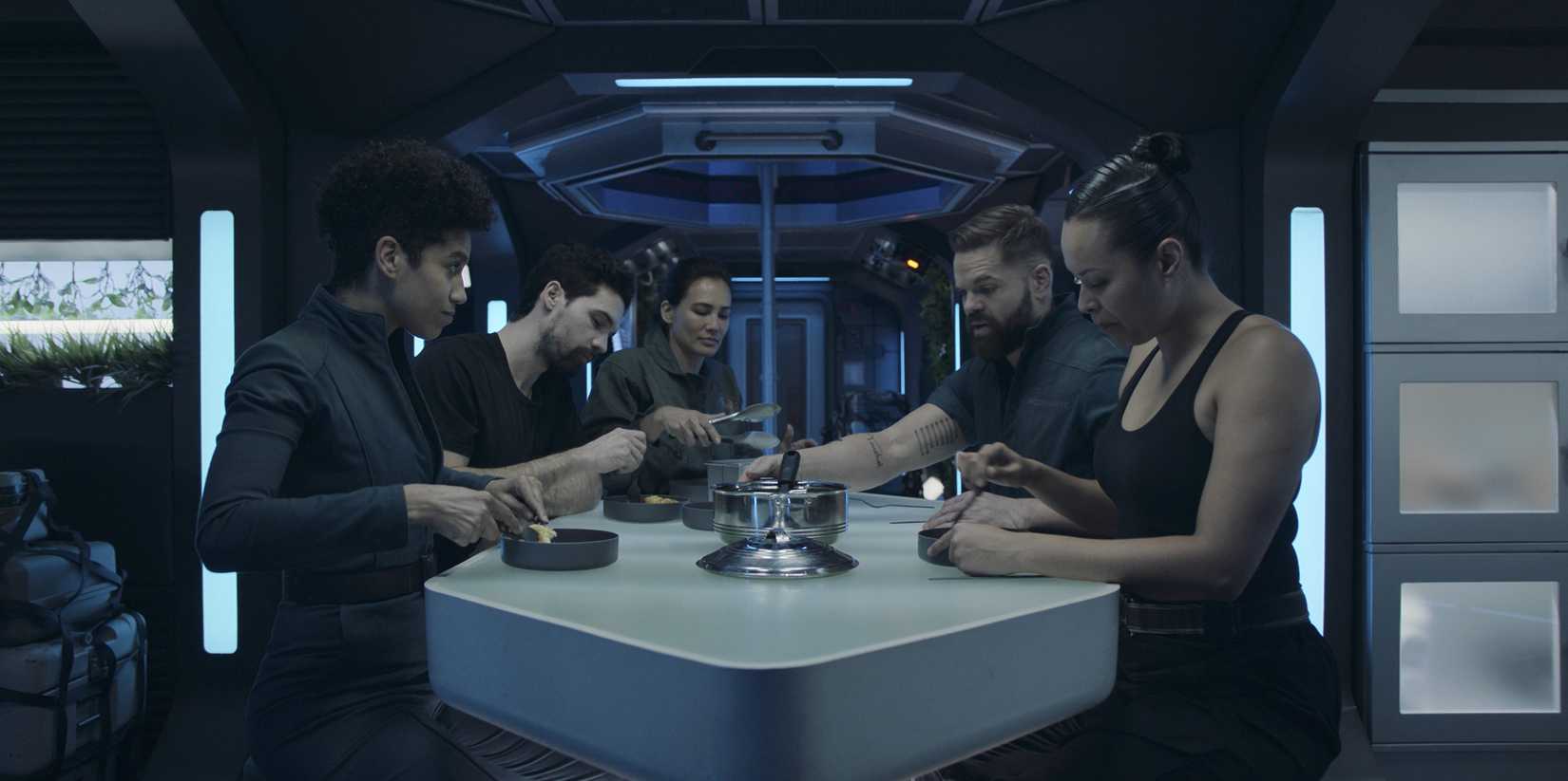 Five spaceship crew members gather around a futuristic table under blue lighting in season 6 of The Expanse
