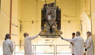 Staff in white lab coats observe a black and gray spacecraft in a room with warm, yellow light.