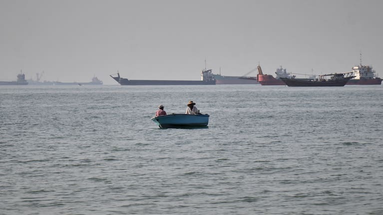 Two men sit in a small boat on the water as a mix of bulk carriers, cargo ships, and service vessels line the horizon in the Strait of Hormuz off Bandar Abbas, Iran, Monday, April 27, 2026.