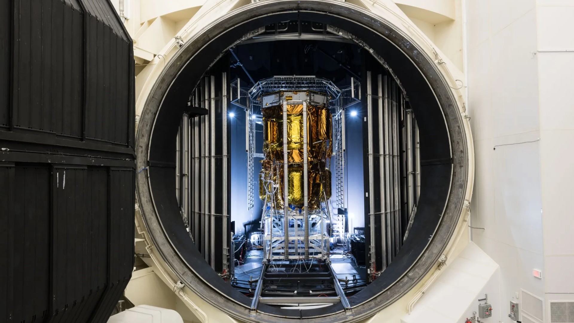 a tall cylindrical spacecraft wrapped in gold foil can be seen through a round door in a large metal chamber