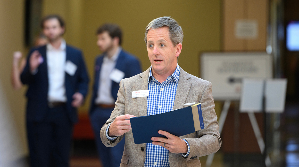 A man reads from a blue folder in front of a group of people.