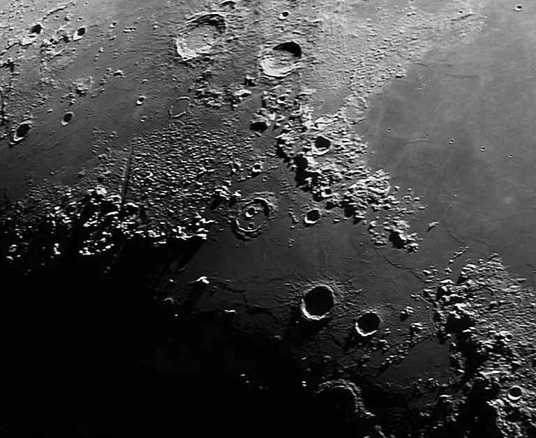 A high-resolution black and white telescopic photograph of the Moon's surface showing the Vallis Alpes (Alpine Valley), a long, straight rille cutting diagonally through the rugged Lunar Alps mountain range. Several craters of varying sizes are visible across the heavily textured terrain, with the terminator line casting deep shadows across the left portion of the image.