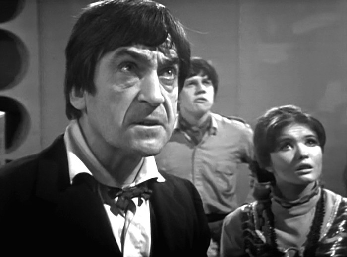web-of-fear Black-and-white Doctor Who scene inside the TARDIS showing the Second Doctor in Victorian-style attire in the foreground, with companions Jamie and Victoria behind him, all looking upward with tense, alarmed expressions.