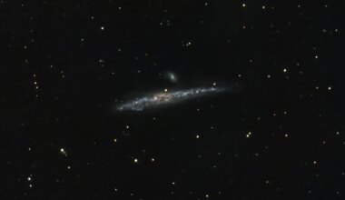 A telescopic photograph of the Whale Galaxy (NGC 4631), an edge-on spiral galaxy appearing as a long, thin streak of blue and white light with a bright, mottled core, set against a black sky filled with foreground stars. A smaller companion galaxy is visible just above the main galaxy near the center of the image.