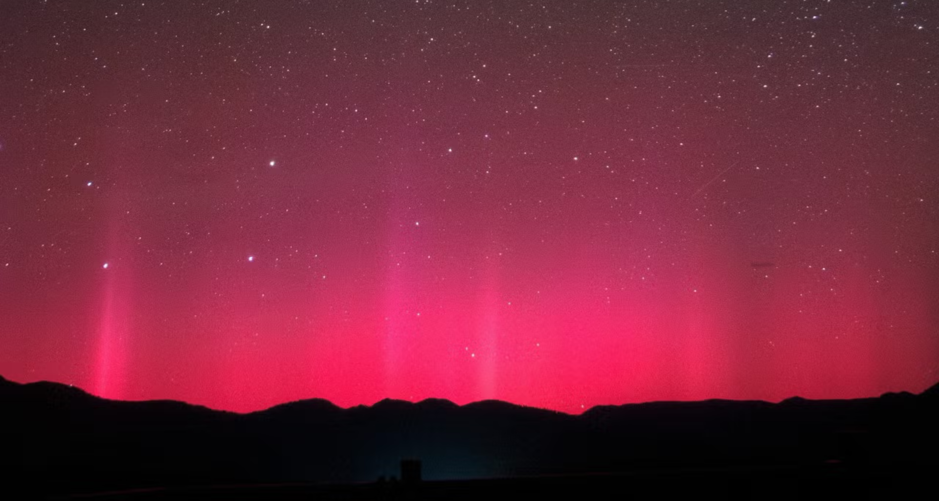 The silhouette of what looks like mountains on the bottom of the screen with bright pink and red lights in the sky above.
