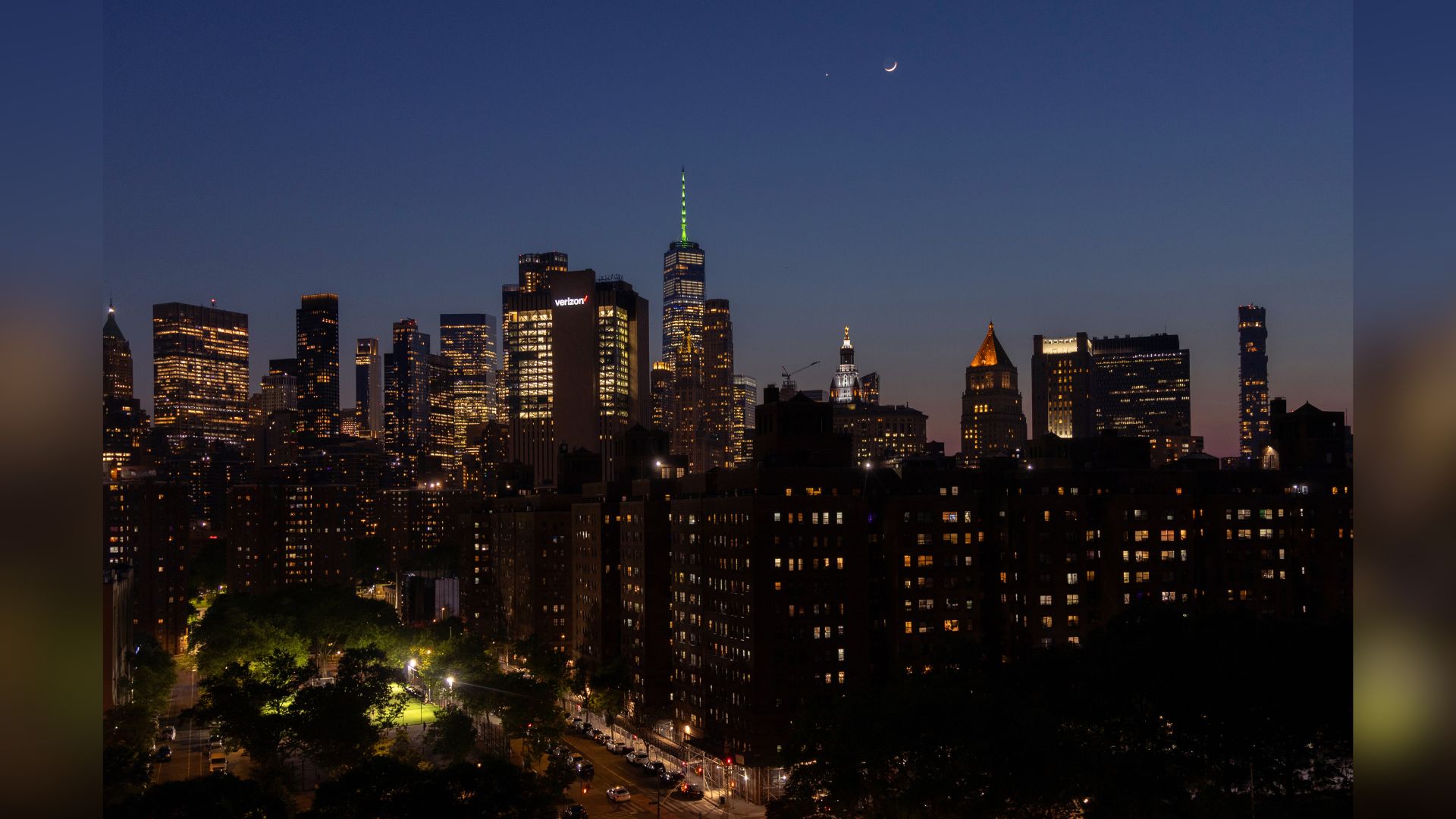 A crescent moon is pictured shining above the New York city skyline in a dark evening sky with a bright planet to its left.