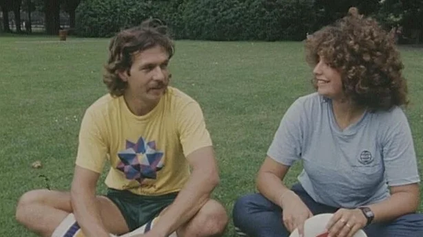 A curly-haired smiling man and a curly-haired smiling woman sitting on the grass