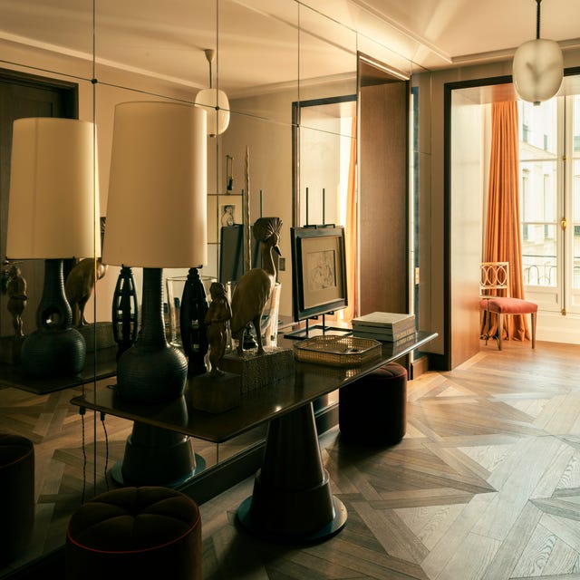Modern room interior with mirrored wall, wooden floor, sculptures, lamps, and window with orange curtains.