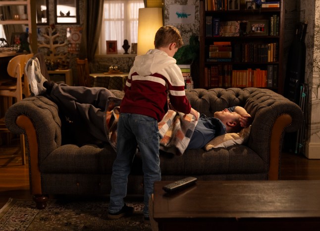 Bertie drapes a blanket over Daniel, who has passed out on the sofa in Corrie