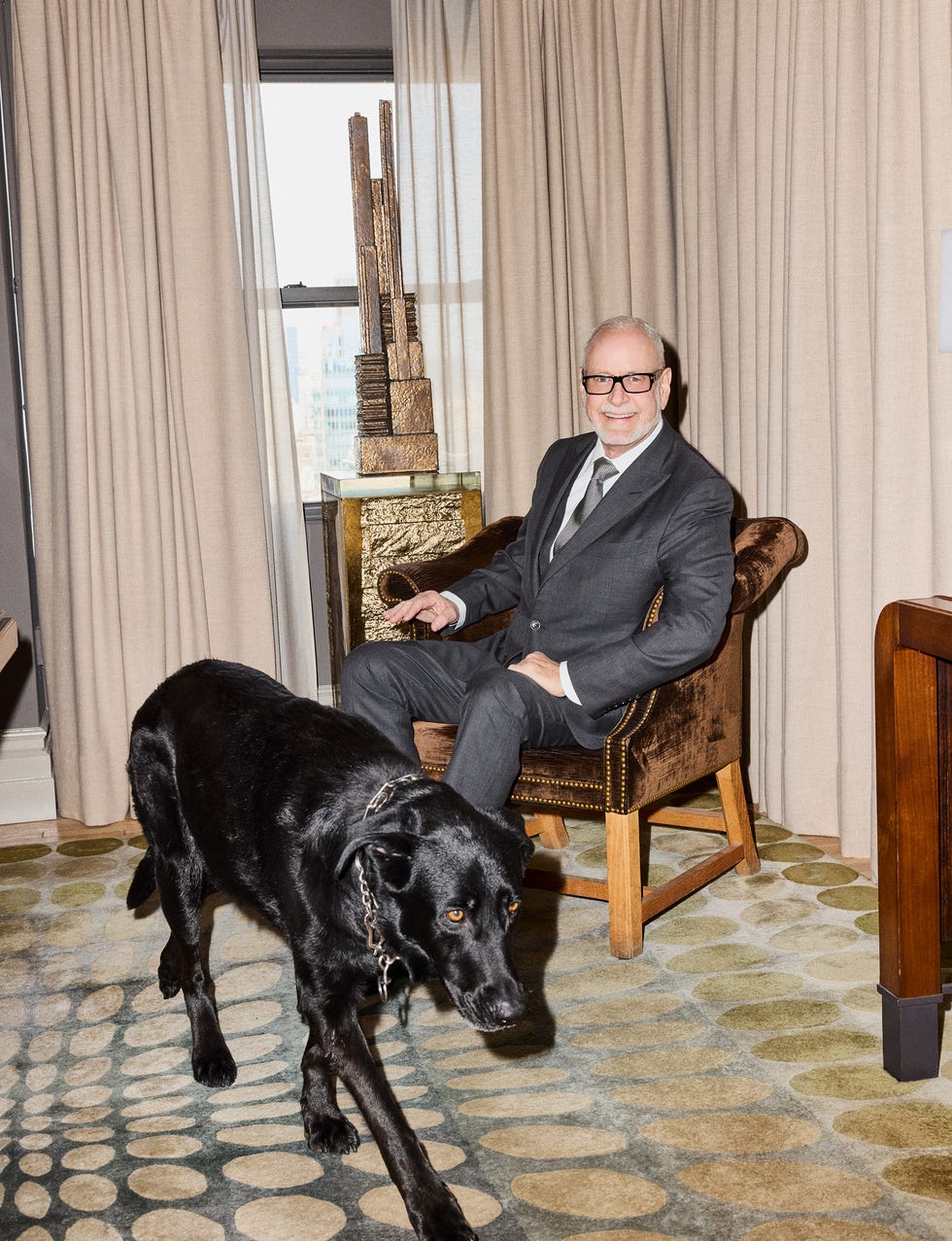 Man in suit sitting in a chair with a large black dog walking in front of him.