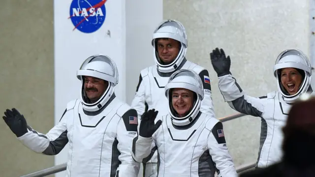 four astronauts in white space suits smiling and waving