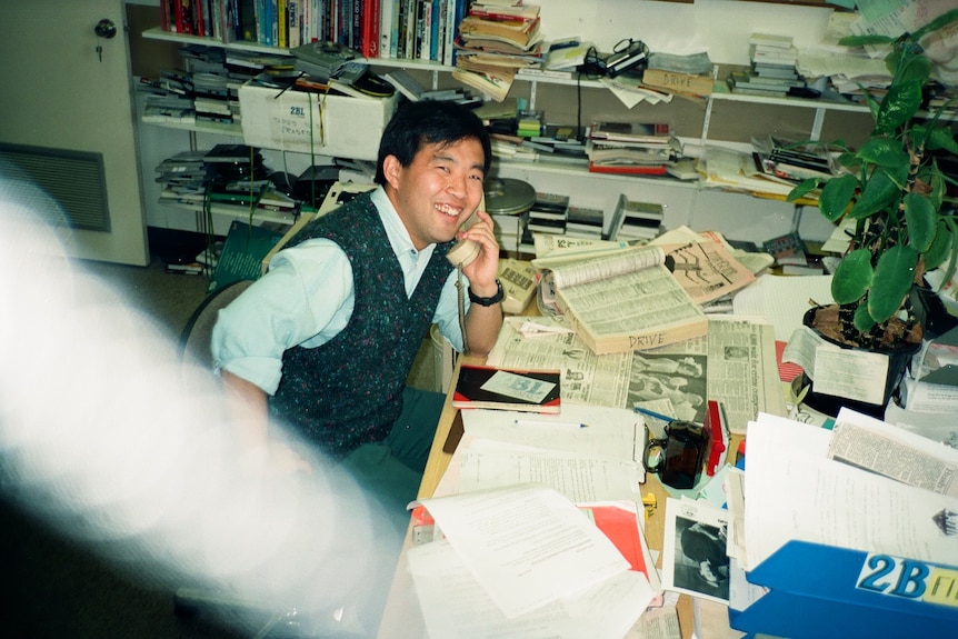 a man smiling while using a landline at a desk covered in newspapers
