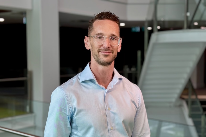 Bespectacled man wearing light blue shirt standing in a foyer with stairs in the background