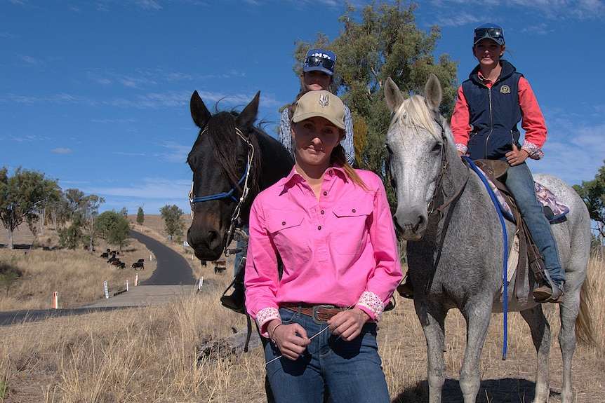 A woman in a pink shirt stands in front of two girls on horses.