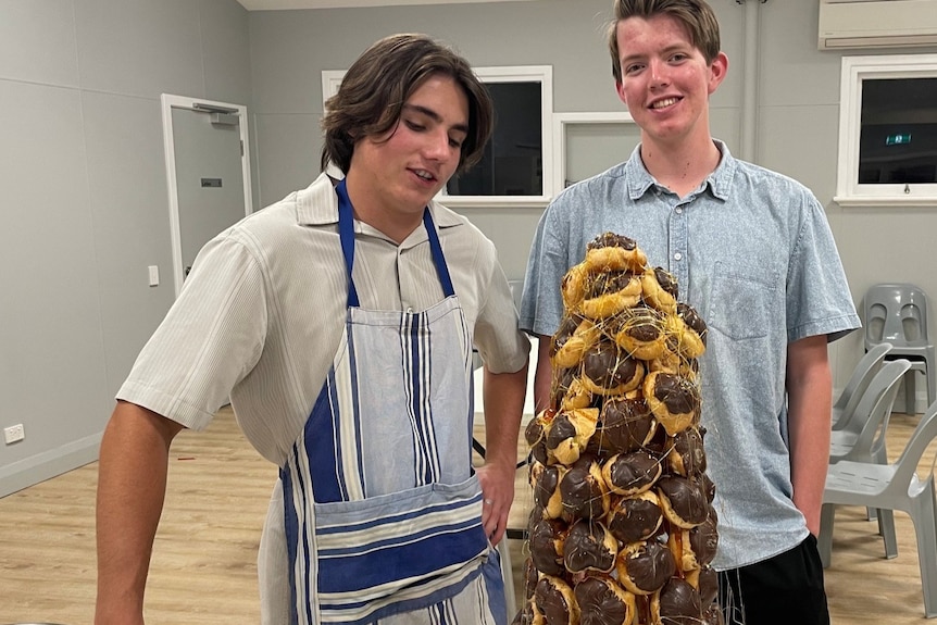 Luke wears an apron and stands next to a male friend. On the table is a tower of profiteroles.
