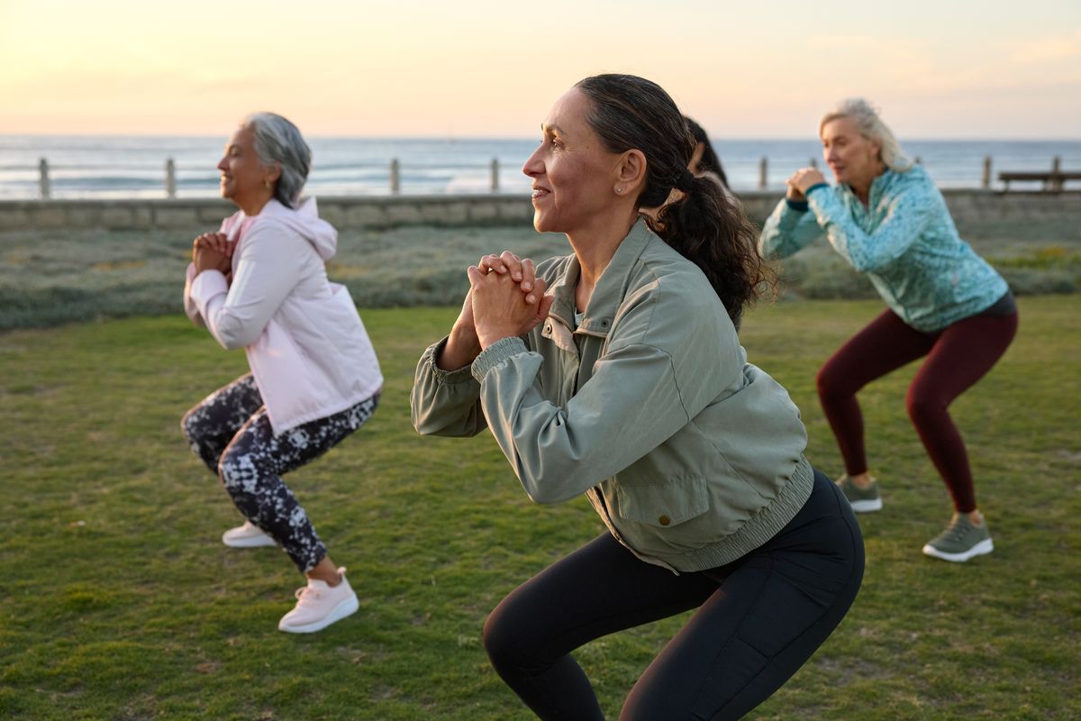 Smiling woman doing exercise with friends in garden. Active female is squatting during morning routine. She is wearing sports clothing.