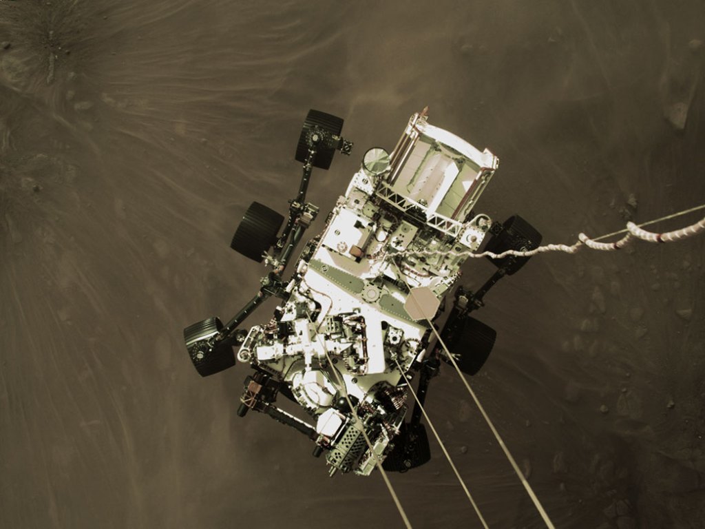 The Mars 2020 descent stage lowers NASA’s Perseverance rover. The six-wheeled rover is seen suspended by cables, with the surface of Mars below. 