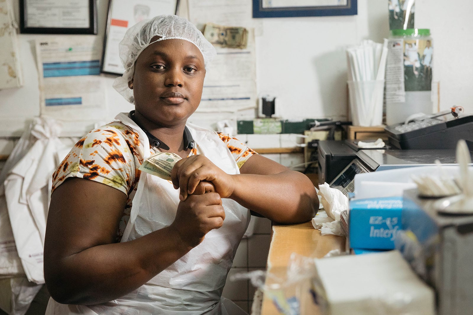 A woman wearing a hair net and apron sits at a counter, holding dollar bills. Behind her are papers, lab coats, and various office supplies, suggesting she is in a small business or restaurant setting.
