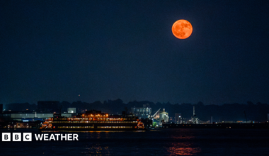 An orange-hued moon hangs in the sky over a river illuminated by lights from nearby properties