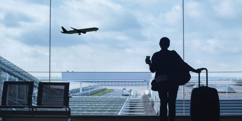 Person in an airport holding a drink and a suitcase with a plane in the background.