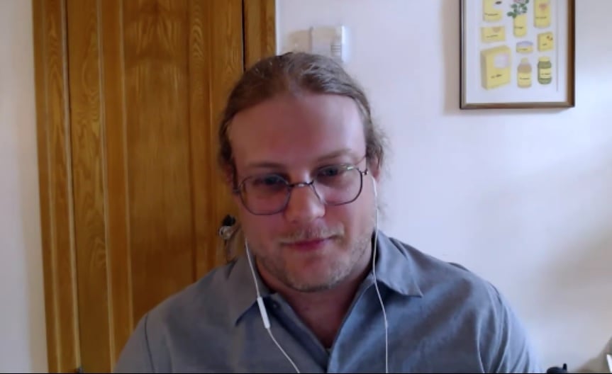 A man with headphones, longer blond hair, glasses in a grey shirt sits in a video call.