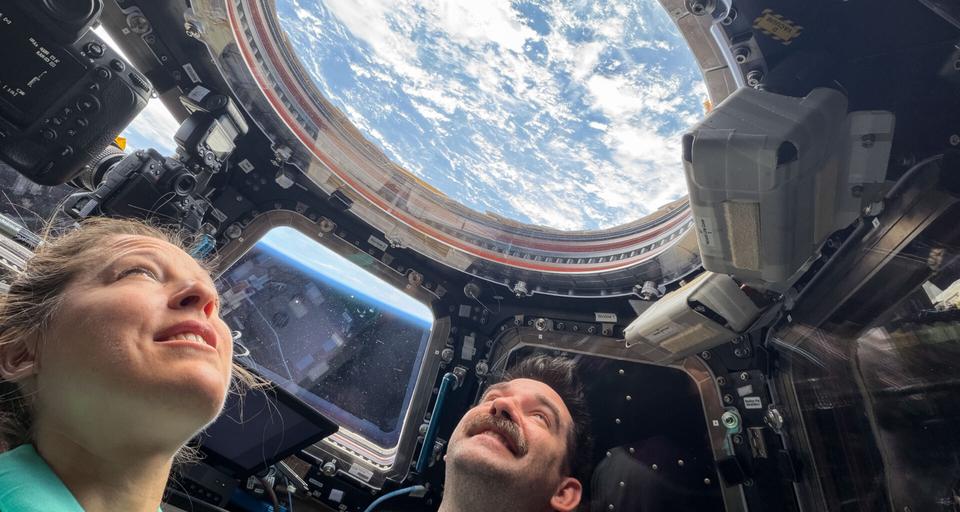 Two astronauts look up through a round window in the International Space Station's cupola. Earth's blue water and white clouds are visible. In a trapezoid-shaped window between the two astronauts, we can see the blue haze of Earth's atmosphere.