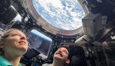 Two astronauts look up through a round window in the International Space Station's cupola. Earth's blue water and white clouds are visible. In a trapezoid-shaped window between the two astronauts, we can see the blue haze of Earth's atmosphere.
