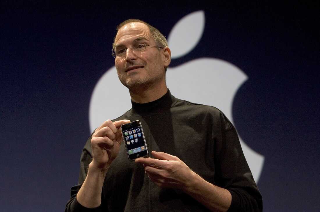 In this 2007 photo, Steve Jobs, then Apple's CEO, holds an iPhone during the smartphone's unveiling. Jobs is wearing a black turtleneck shirt, and he's standing in front of a black backdrop that's displaying the Apple logo in white.